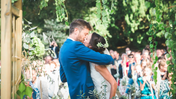 Photographe de mariage saisit un baiser passionné entre les mariés sous une arche verdoyante à Bordeaux.
