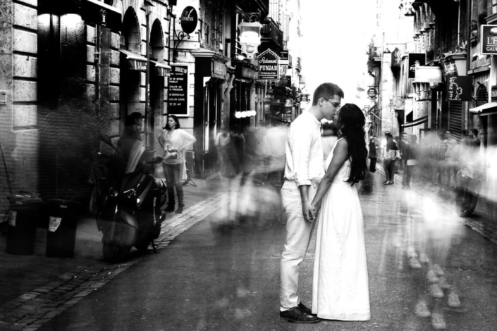 Couple s'embrassant tendrement dans une rue animée de Bordeaux, en noir et blanc, capturé par La Focale d'Olga, photographe de mariage.