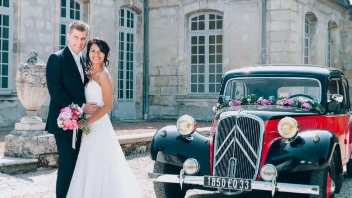Mariés souriants posant à côté d'une voiture classique décorée de fleurs, photographiés par La Focale d'Olga, offrant des services accessibles de photographie de mariage à Bordeaux, Gironde et dans le Sud-Ouest.