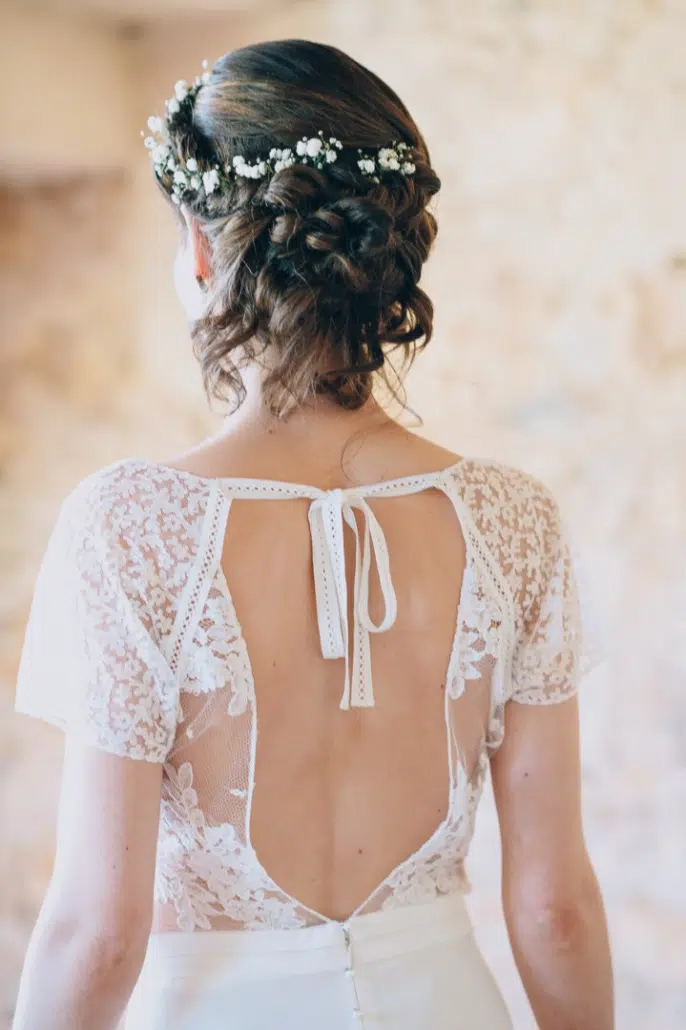 Vue arrière d'une mariée révélant une coiffure complexe ornée de fleurs blanches et une robe en dentelle délicate, capturée en Gironde par lafocaledolga.