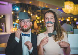 Couple s'amusant avec des accessoires de photomaton lors d'une soirée de mariage, décor festif en arrière-plan.