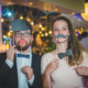 Couple s'amusant avec des accessoires de photomaton lors d'une soirée de mariage, décor festif en arrière-plan.