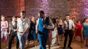 Invités dansant joyeusement lors d'une soirée de mariage dans une salle aux murs de pierre illuminée par des lumières colorées.